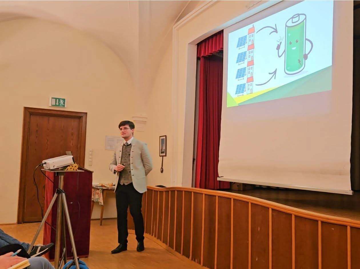 Ein Mann im Anzug hält eine Präsentation in einem Raum mit einer Projektionsleinwand, die ein Diagramm einer Batterie und Solarmodule zeigt.