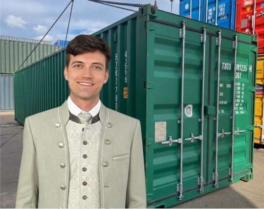 A man in formal attire stands in front of a large green shipping container, smiling.