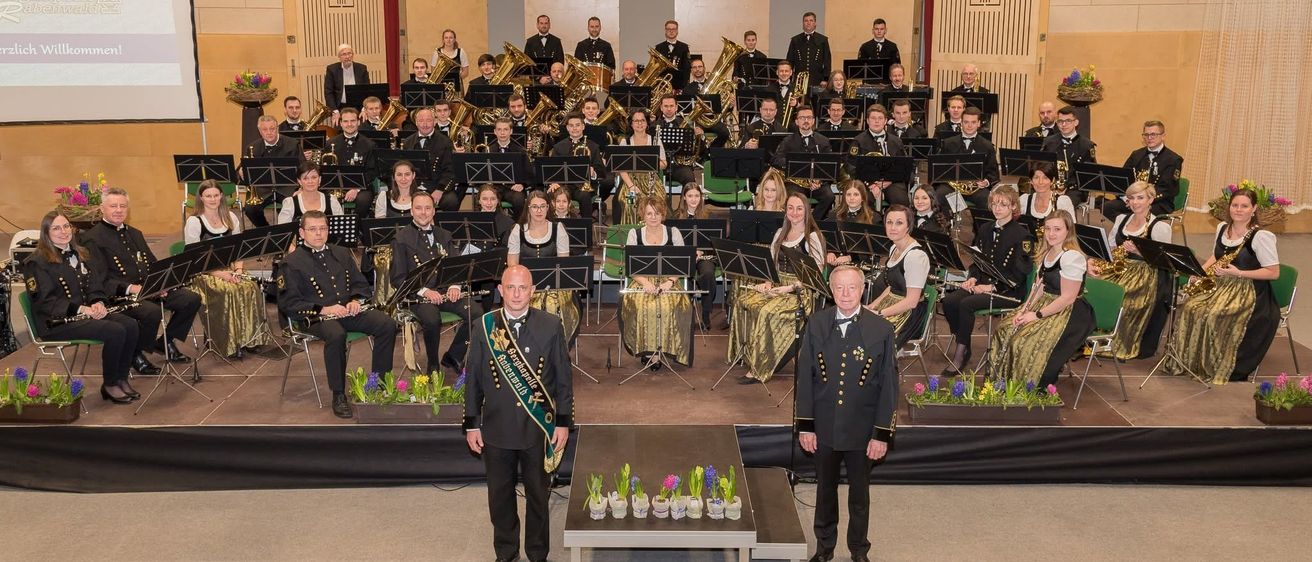 Eine große Gruppe von Musikern in formeller Kleidung, einschließlich Männer und Frauen, steht auf einer Bühne mit Musikinstrumenten, wahrscheinlich für eine Aufführung. Zwei Männer vorne tragen Schärpen.