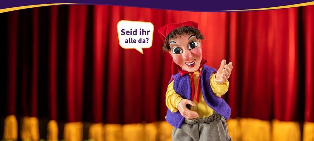 Eine Puppe mit rotem Hut steht auf der Bühne, mit einem roten Vorhang dahinter. Sie scheint an das Publikum zu sprechen.