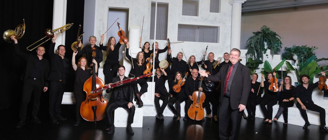 Eine Gruppe von Musikern, sowohl sitzend als auch stehend, posiert für ein Foto. Sie halten verschiedene Instrumente wie Violinen, Celli und ein Fagott. Ein Mann in der Mitte hält eine Trompete. Der Hintergrund zeigt eine weiße Struktur mit Fenstern.