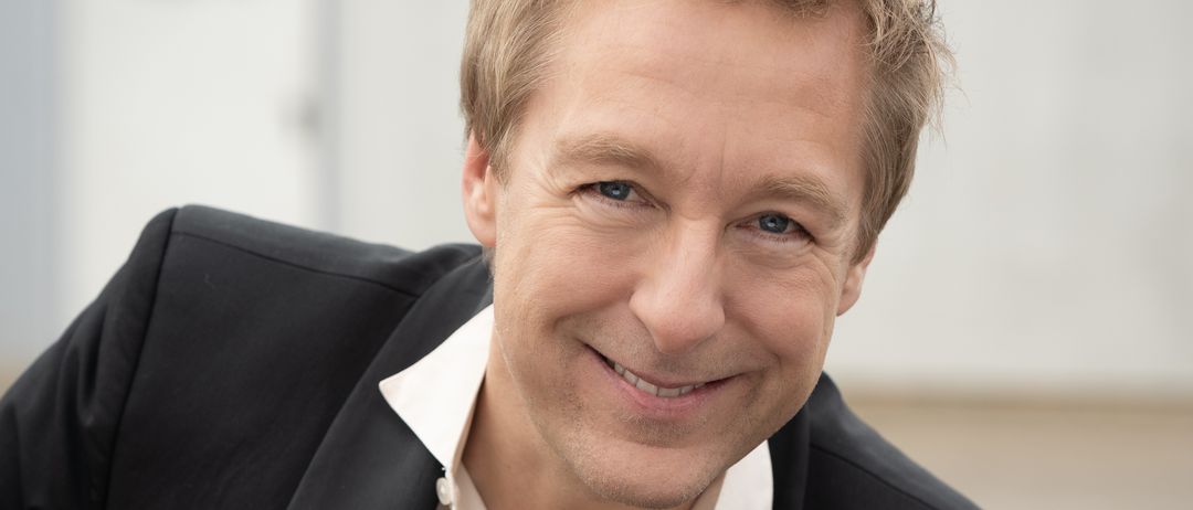 Ein lächelnder Mann mit kurzen blonden Haaren in einem schwarzen Anzug und weißem Hemd posiert für ein Foto.