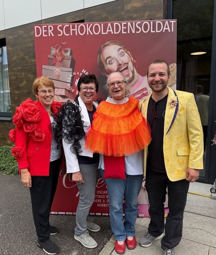 Vier Personen stehen vor einem großen Werbeplakat mit einer Person in einem gelben Jackett. Sie alle lächeln und posieren für ein Foto.