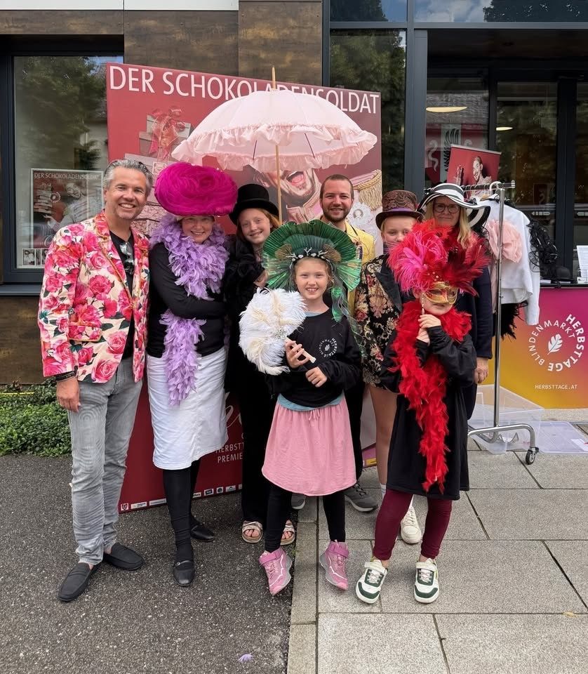 Eine Gruppe von Menschen in farbenfrohen Kostümen posiert für ein Foto vor einem roten Banner mit Text, einem Regenschirm und einem Gebäude im Hintergrund.
