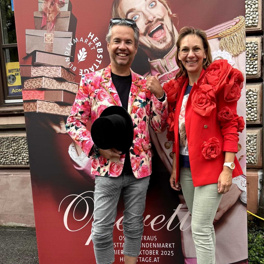 Ein Paar steht vor einem roten Plakat mit einem großen Bild eines Mannes in einer goldenen Robe. Der Mann im Bild hat einen überraschten Gesichtsausdruck. Der Mann im Vordergrund trägt einen rosafarbenen Blumenanzug und hält einen schwarzen Hut. Die Frau trägt einen roten Blazer mit Blumendetails.
