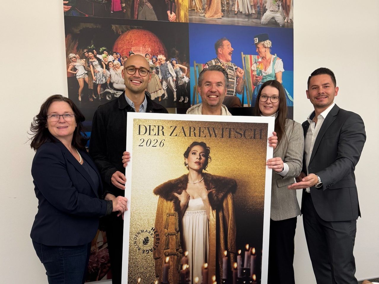 Fünf Personen stehen hinter einem großen Poster einer Frau. Die Frau trägt einen Pelzmantel und hält Kerzen. Das Poster lautet 'Der Zarewitsch 2026'.