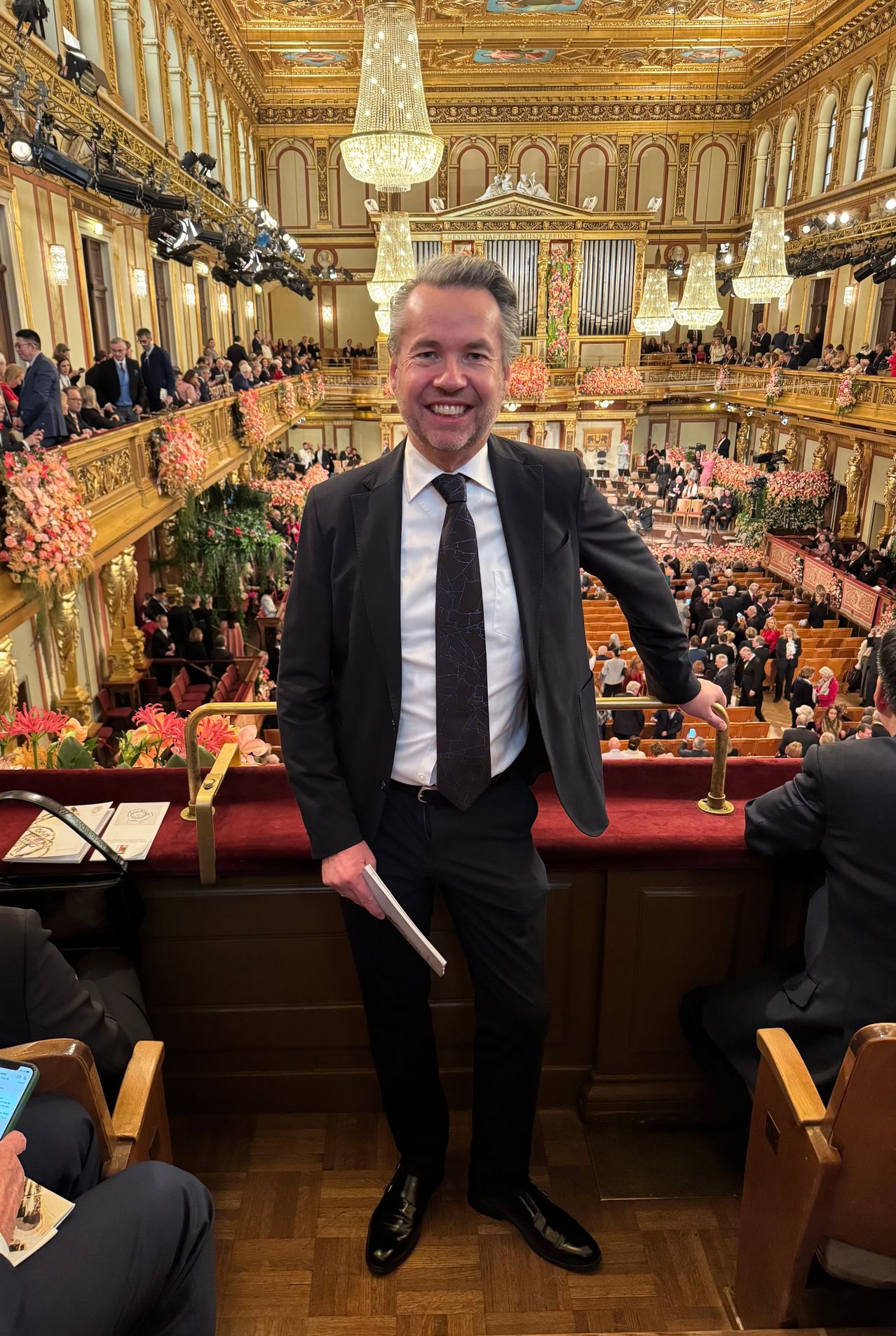 Ein Mann in einem Anzug steht lächelnd in einem großen Auditorium. Hinter ihm sitzt ein Publikum, und der Raum ist mit Blumen geschmückt.