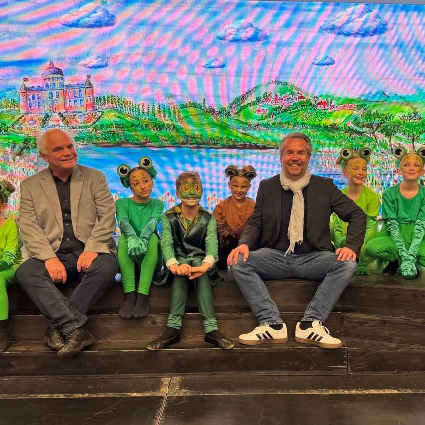Zwei Männer und mehrere Kinder in grünen Kostümen sitzen auf einer Bühne mit buntem Hintergrund. Sie scheinen ein Theaterstück aufzuführen.