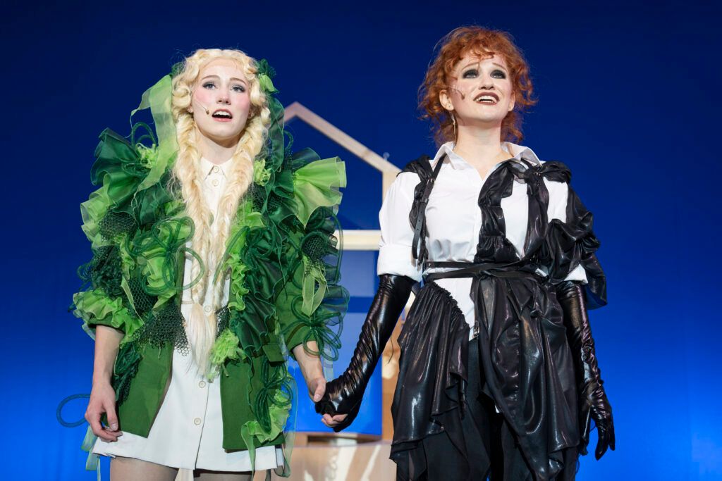 Zwei Frauen auf der Bühne, eine in Grün mit einem Mikrofon, die andere in Schwarz, halten Händchen. Sie sind wahrscheinlich bei einer Aufführung. Der Hintergrund ist blau mit einem weißen Hausumriss.