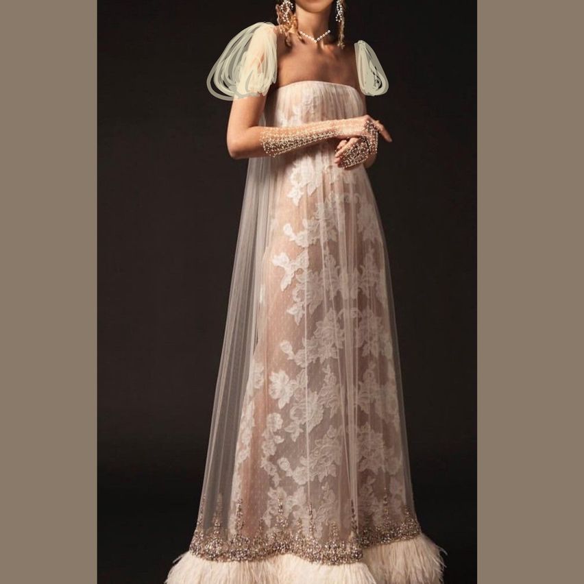 Eine Frau in einem langen, eleganten Kleid mit aufwendiger Spitze und Federdetails, vor einem dunklen Hintergrund stehend.