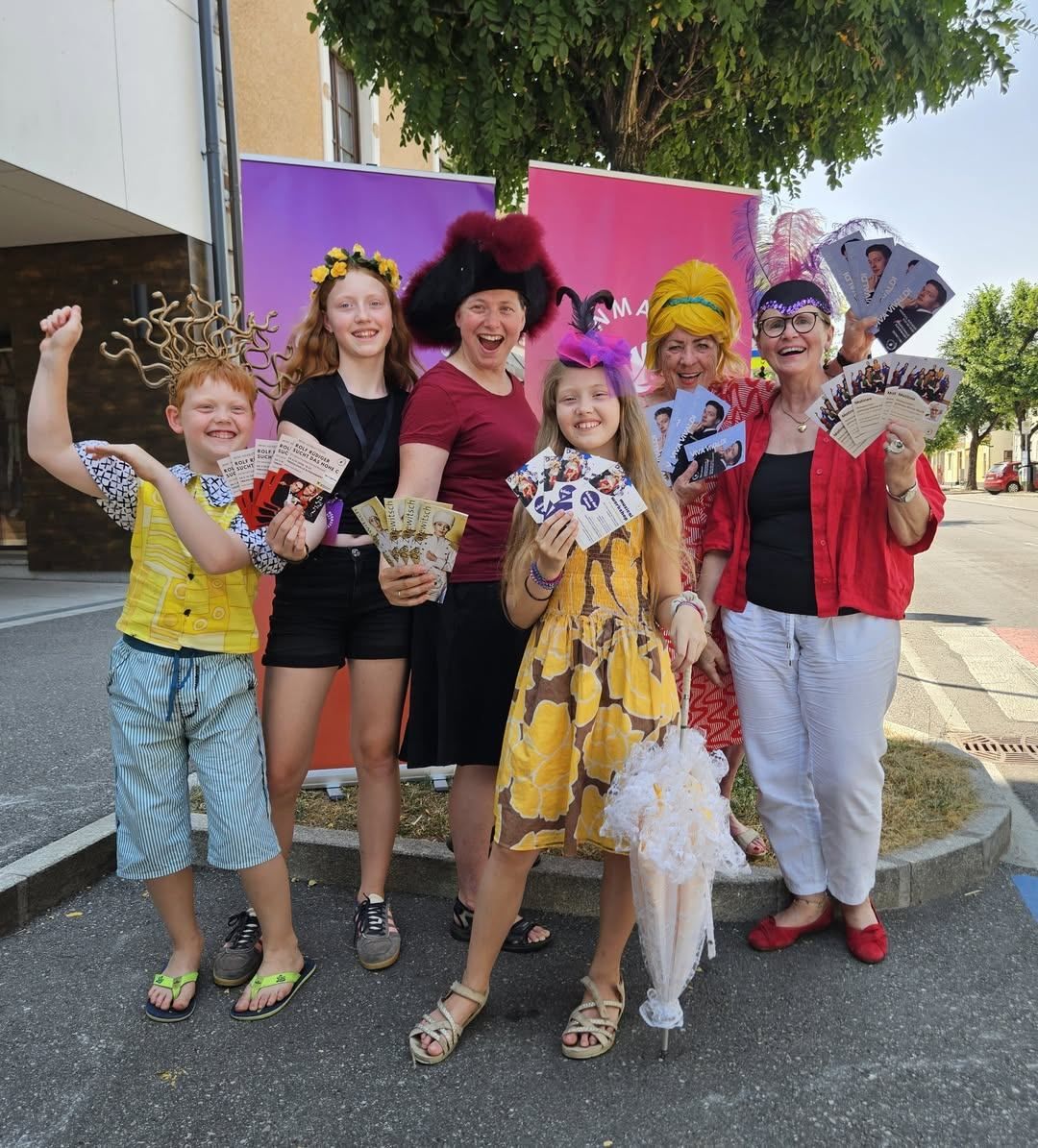 Eine Gruppe von Menschen, darunter Kinder und Erwachsene, posiert für ein Foto, während sie verschiedene Hüte tragen und Eintrittskarten halten. Sie stehen vor einem Gebäude mit einem rosa Banner.