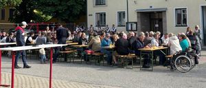 Eine Gruppe von Menschen sitzt um Tische herum draußen in einem Innenhof. Bäume, ein Gebäude und ein Rollstuhl sind in der Szene zu sehen.
