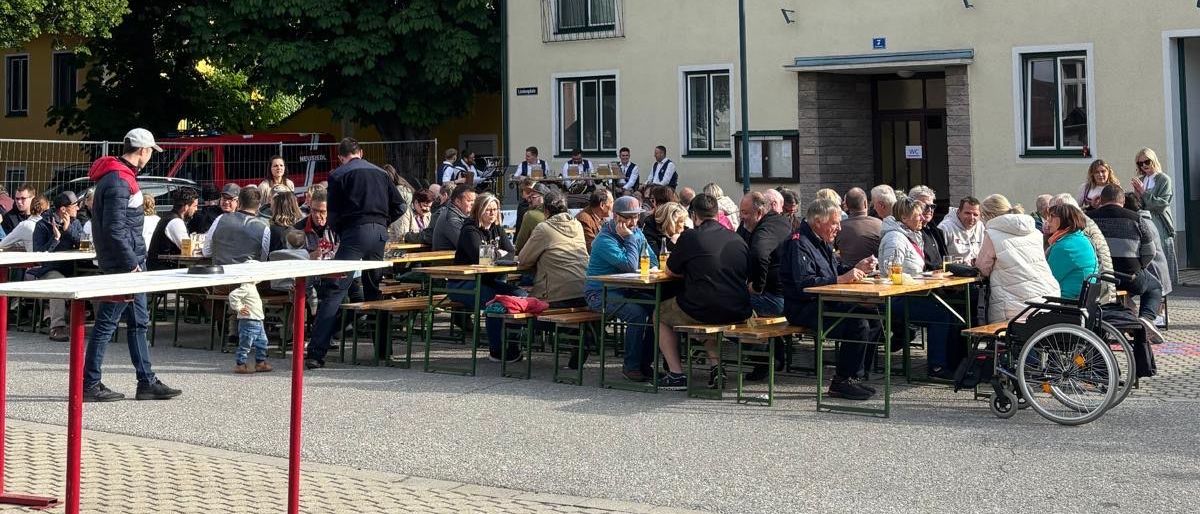 Eine Gruppe von Menschen sitzt um Tische herum draußen in einem Innenhof. Bäume, ein Gebäude und ein Rollstuhl sind in der Szene zu sehen.