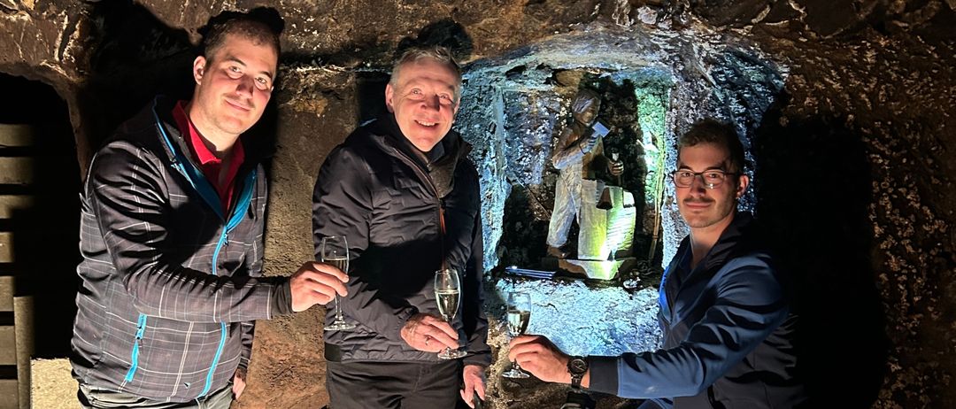 Drei Männer in Jacken und Hosen stehen in einer Höhle und halten Weingläser in den Händen. Sie lächeln und scheinen für ein Foto zu posieren.