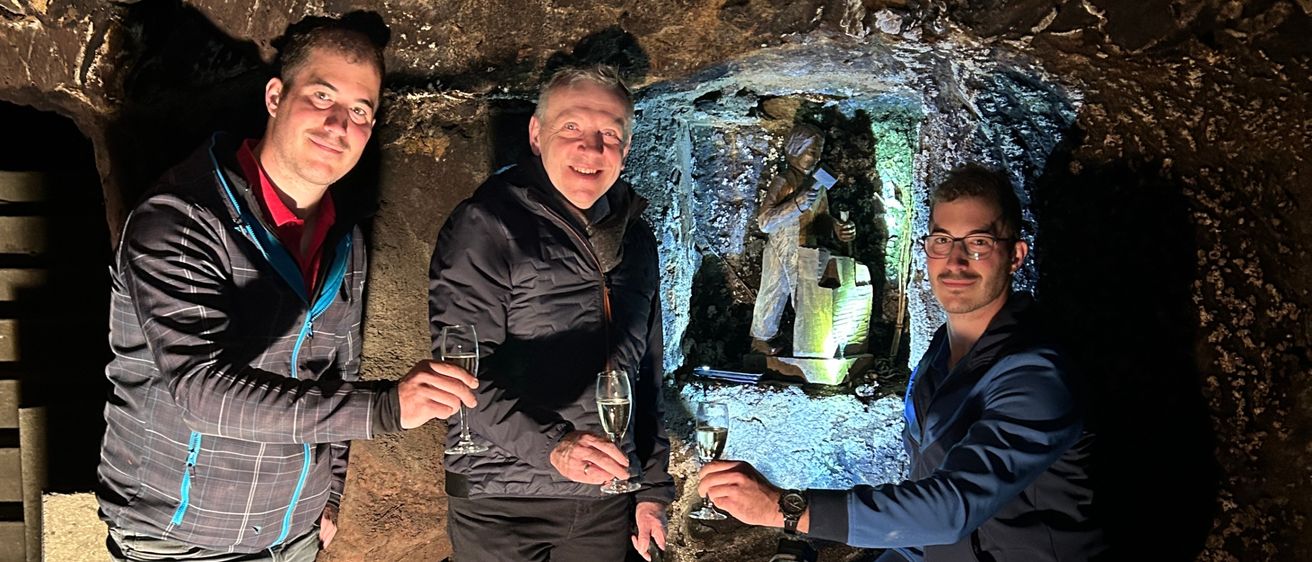 Drei Männer in Jacken und Hosen stehen in einer Höhle und halten Weingläser in den Händen. Sie lächeln und scheinen für ein Foto zu posieren.