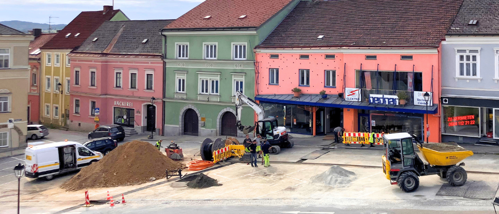 Eine Baustelle mit einem Haufen Erde, einem gelben Traktor und einem weißen Bagger. Die Gebäude sind in verschiedenen Farben gestrichen, darunter pink, grün und braun. Eines der Gebäude hat einen blauen Vorbau. Arbeiter sind anwesend, und es gibt Schilder an den Gebäuden.