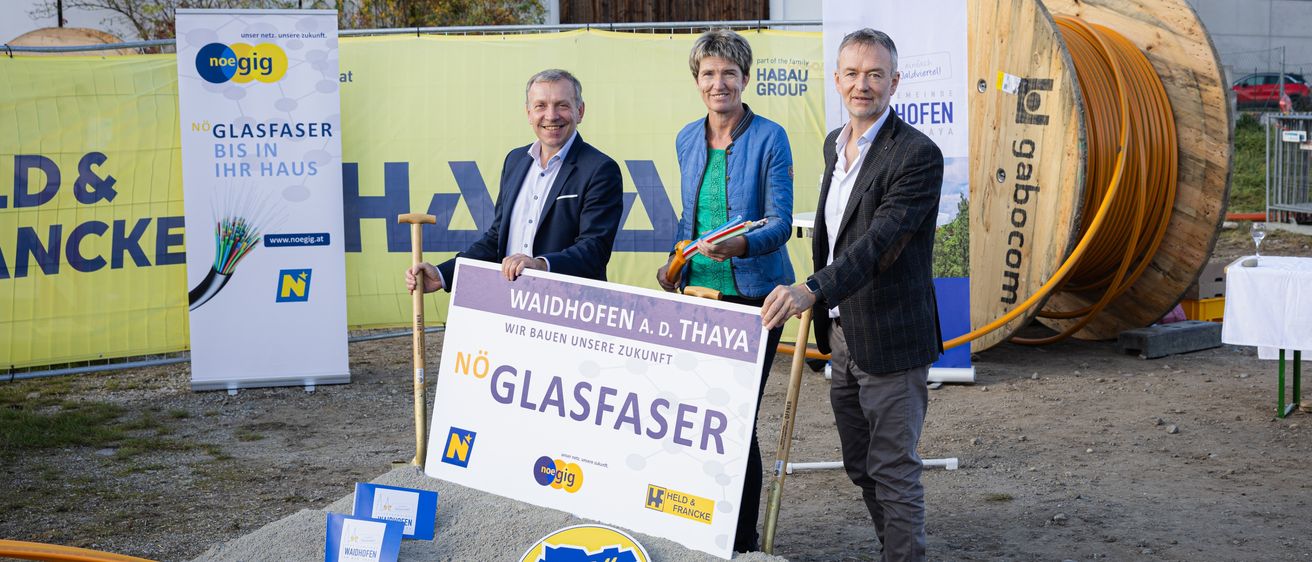 Drei Personen stehen vor einem Schild mit der Aufschrift Waidhofen. Die beiden Männer halten Schaufeln, und die Frau hält ein Bündel Drähte. Dahinter befindet sich ein Banner mit dem Text HAH und einem Logo.