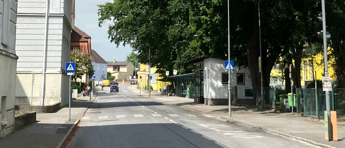 Eine ruhige Straße mit weißen Gebäuden auf beiden Seiten, gesäumt von Bäumen und Straßenlaternen. Ein Auto ist rechts geparkt.