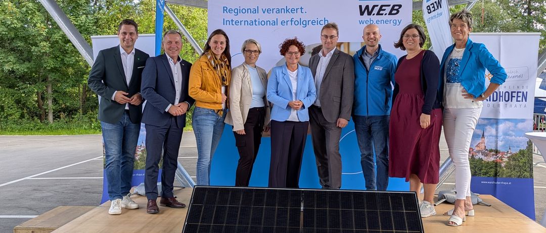 Eine Gruppe von Menschen steht vor einem Solarpanel und einem Banner mit der Aufschrift 'Regional verankert. International erfolgreich.' Sie sind alle formell gekleidet.