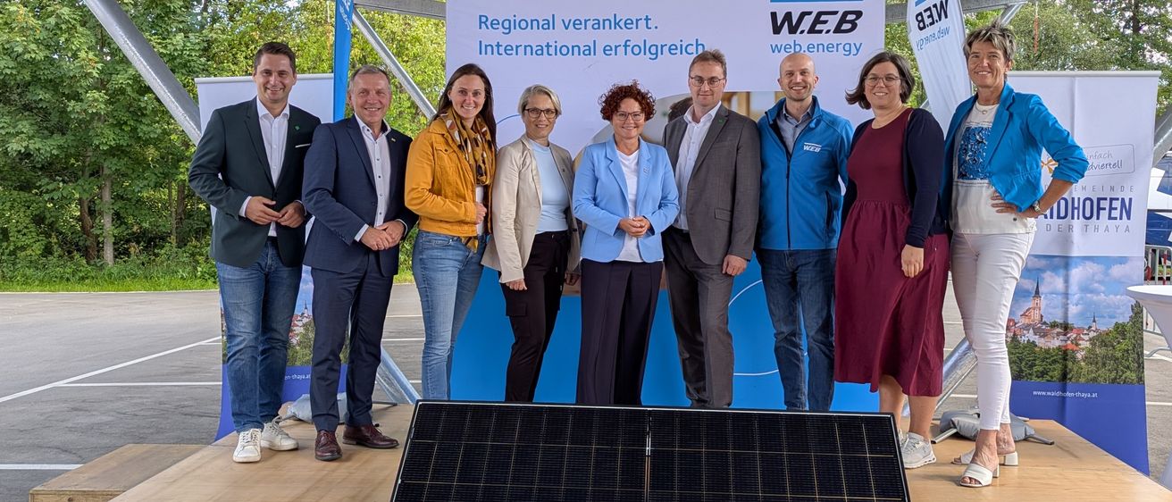 Eine Gruppe von Menschen steht vor einem Solarpanel und einem Banner mit der Aufschrift 'Regional verankert. International erfolgreich.' Sie sind alle formell gekleidet.
