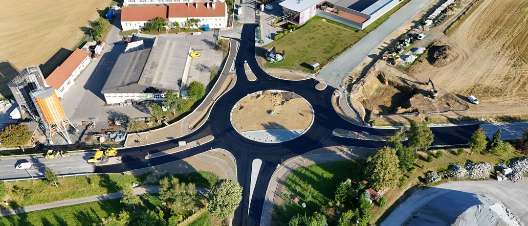 Ein Luftbild einer Baustelle mit einem kreisförmigen Straßenkreuz, neu asphaltierten Straßen, umgeben von Gebäuden, Bäumen und offenen Rasenflächen.