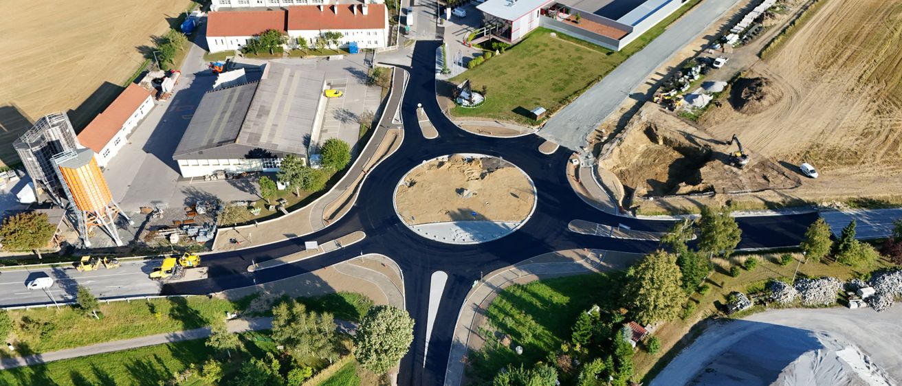 Ein Luftbild einer Baustelle mit einem kreisförmigen Straßenkreuz, neu asphaltierten Straßen, umgeben von Gebäuden, Bäumen und offenen Rasenflächen.