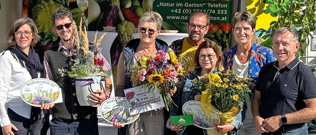 Eine Gruppe von Menschen steht vor einem Stand, Blumen und Urkunden in der Hand. Sie sind lässig gekleidet, einige tragen Brillen. Hinter ihnen zeigt ein Banner Gemüse und Kontaktinformationen.