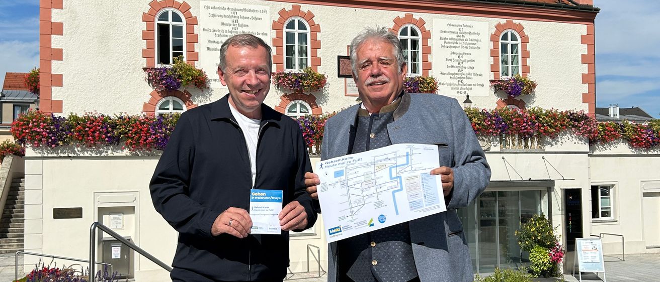 Zwei Männer stehen vor einem Gebäude mit rotem Dach und Turm. Sie halten ein Blatt Papier mit Zeichnungen in der Hand und lächeln.