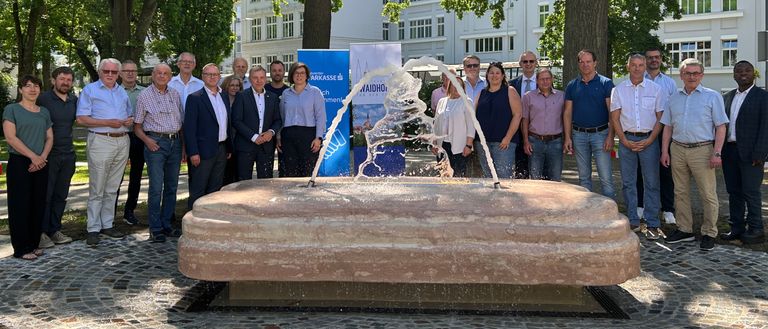 Eine Gruppe von Menschen, einige in Geschäftskleidung, posiert für ein Foto um eine Wasserfontäne in einem Park mit Bäumen und einem weißen Gebäude im Hintergrund.