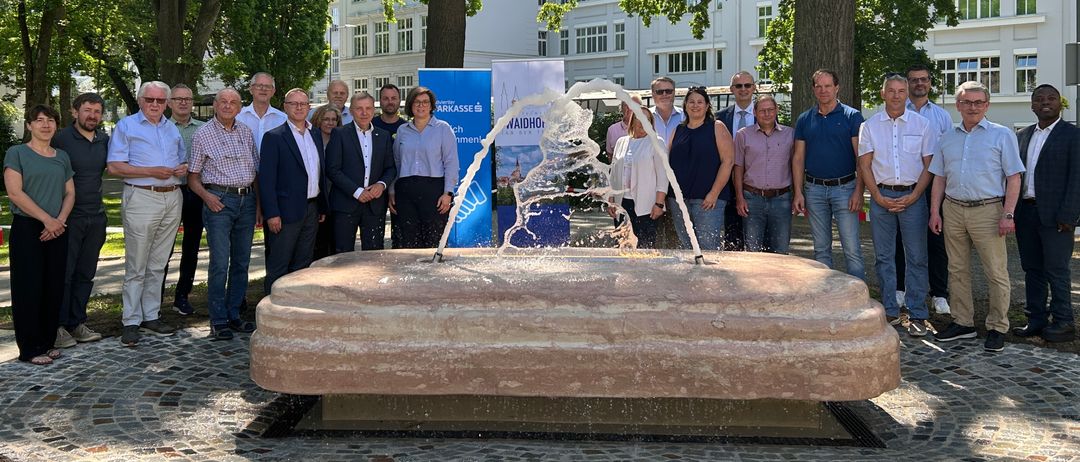 Eine Gruppe von Menschen, einige in Geschäftskleidung, posiert für ein Foto um eine Wasserfontäne in einem Park mit Bäumen und einem weißen Gebäude im Hintergrund.