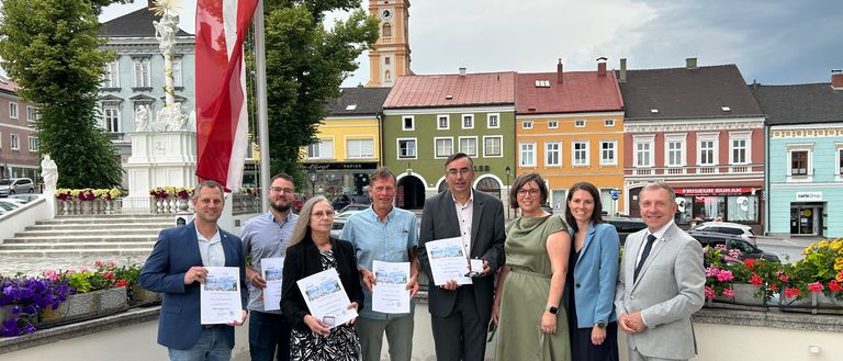 Eine Gruppe von Menschen in formeller Kleidung, die Urkunden halten, posiert für ein Foto vor einem Stadtplatz mit Gebäuden und einer Fahne.