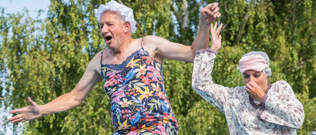 Ein älteres Paar, der Mann in einem Blumenkleid und die Frau in einem Blumengewand, springt gemeinsam von einer Sprungbrett, umgeben von grünen Bäumen.