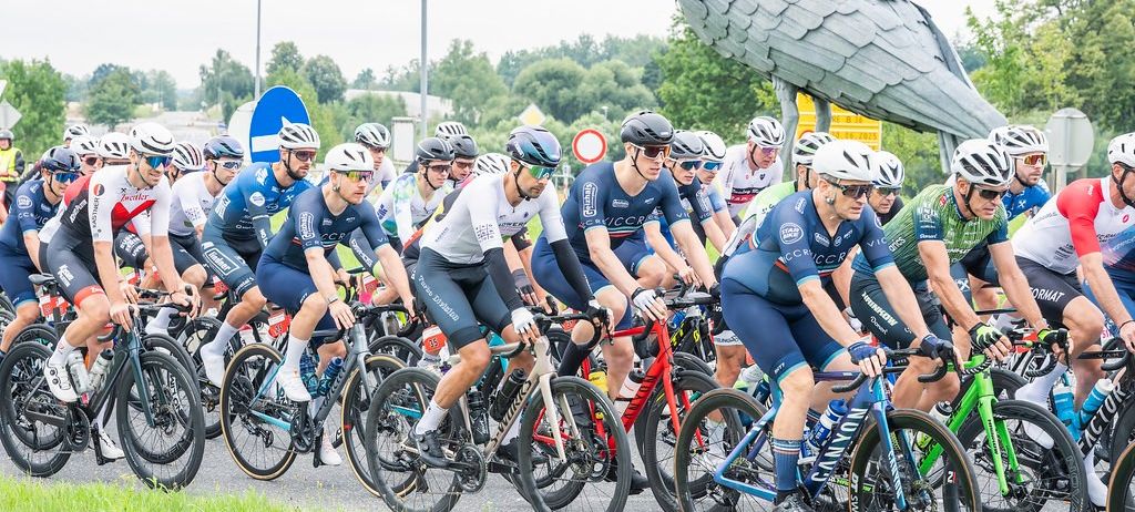 Eine Gruppe von Radfahrern in übereinstimmenden blauen Uniformen fährt gemeinsam auf einer Straße, mit einer großen Vogelskulptur im Hintergrund.