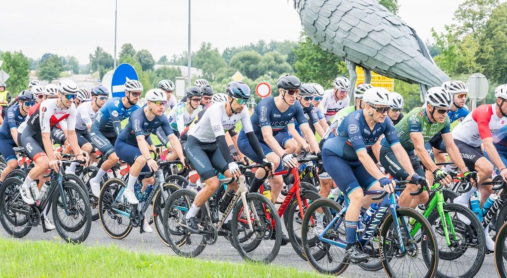 Eine Gruppe von Radfahrern in übereinstimmenden blauen Uniformen fährt gemeinsam auf einer Straße, mit einer großen Vogelskulptur im Hintergrund.