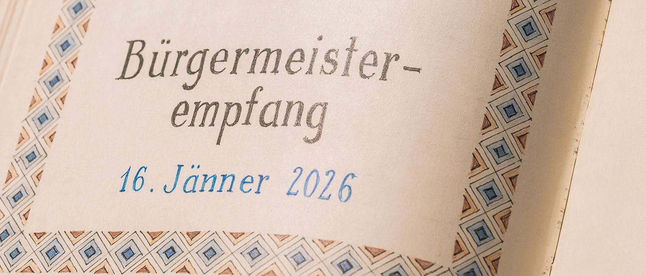 Ein offenes Buch zeigt eine Zeichnung eines Fisches über dem Text 'Bürgermeister-Empfang 16. Janner 2026'. Das Design besteht aus einem blauen und braunen geometrischen Muster.