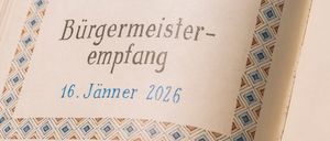 An open book displays a drawing of a fish above the text 'Burgermeister-Empfang 16. Janner 2026'. The design features a blue and brown geometric pattern.