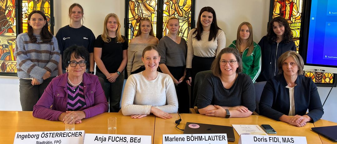 Eine Gruppe von Frauen posiert für ein Foto vor einem Tisch. Sie befinden sich in einem Konferenzraum mit Buntglasfenstern. Der Tisch hat Namensschilder.