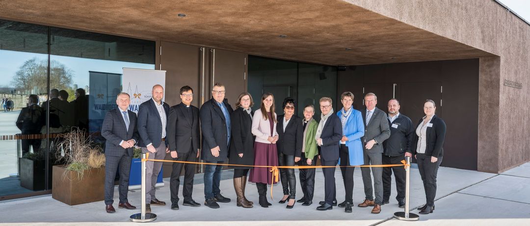 Eine Gruppe von Fachleuten in formeller Kleidung steht vor einem modernen Gebäude. Sie halten ein orangefarbenes Band, das ein Ereignis oder eine Eröffnung symbolisiert.