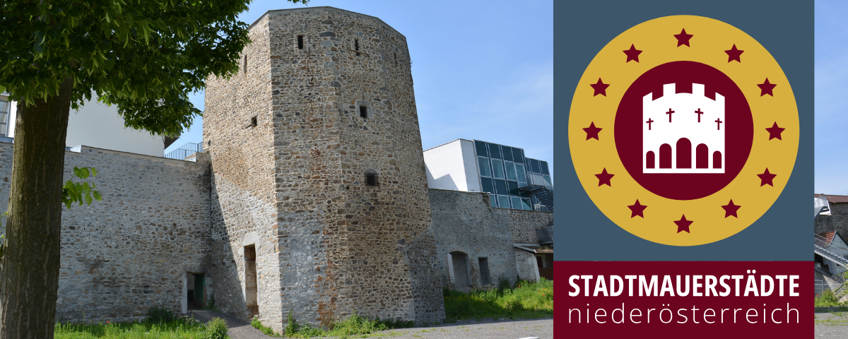 Ein alter Steinturm und ein modernes Gebäude stehen nebeneinander mit einem Grasbereich davor. Der Text 'Stadtmauer niederoess' ist auf einem gelben Hintergrund angezeigt.
