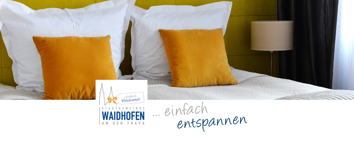 Ein ordentlich gemachtes Bett mit weißen und gelben Kissen. Eine Lampe steht auf dem Nachttisch. Ein blaues Logo lautet 'Waidhofen an der Thaya'.