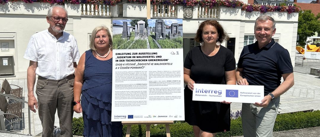 Vier Erwachsene stehen vor einem historischen Gebäude mit einem Banner und einer Ausstellung. Sie halten ein Plakat und scheinen für ein Foto zu posieren. Dahinter ist ein großes Plakat mit Text in einer fremden Sprache zu sehen.