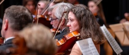 Ein Symphonieorchester tritt auf der Bühne auf. Eine Geigerin spielt die Geige, und der Rest der Musiker spielt ebenfalls ihre Instrumente. Das Publikum beobachtet die Aufführung.