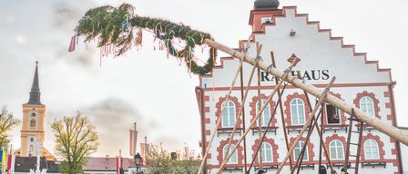 Ein Plakat bewirbt ein traditionelles Maibaumfest in Waidhofen an der Thaya. Feuerwehrleute stellen einen Maibaum vor einem Gebäude auf. Der Text lautet 'Wir freuen uns auf Sie!' und 'Gemeinsames Maibaumaufstellen'. Das Datum ist der 30. April 2026.