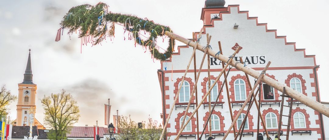 Ein Plakat bewirbt ein traditionelles Maibaumfest in Waidhofen an der Thaya. Feuerwehrleute stellen einen Maibaum vor einem Gebäude auf. Der Text lautet 'Wir freuen uns auf Sie!' und 'Gemeinsames Maibaumaufstellen'. Das Datum ist der 30. April 2026.