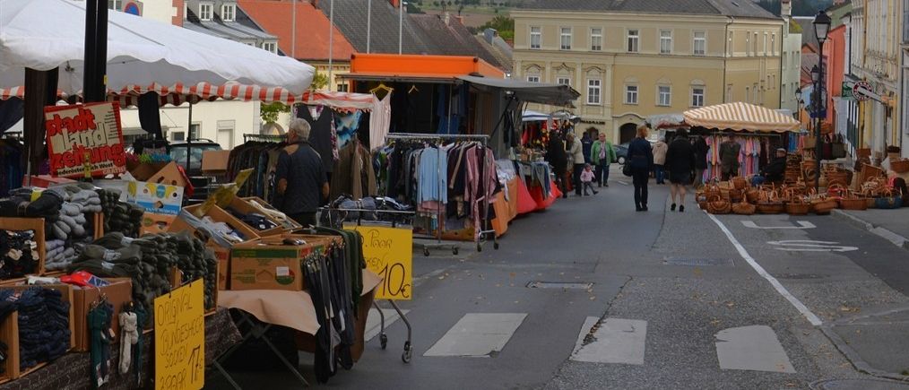 A street market is held on the side of a street. Stalls are set up with various items, including clothes, fruits, and vegetables. There are people walking and shopping on the street. Buildings are on both sides of the street, and mountains are in the distance.