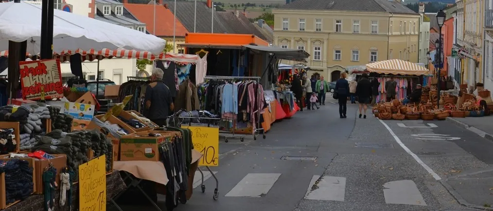 Ein Straßenmarkt mit verschiedenen Ständen, die Kleidung und andere Artikel verkaufen. Die Stände stehen unter weißen und orangefarbenen Zelten. Menschen gehen auf der Straße. Auf beiden Seiten der Straße befinden sich Gebäude. Der Himmel ist blau mit weißen Wolken.