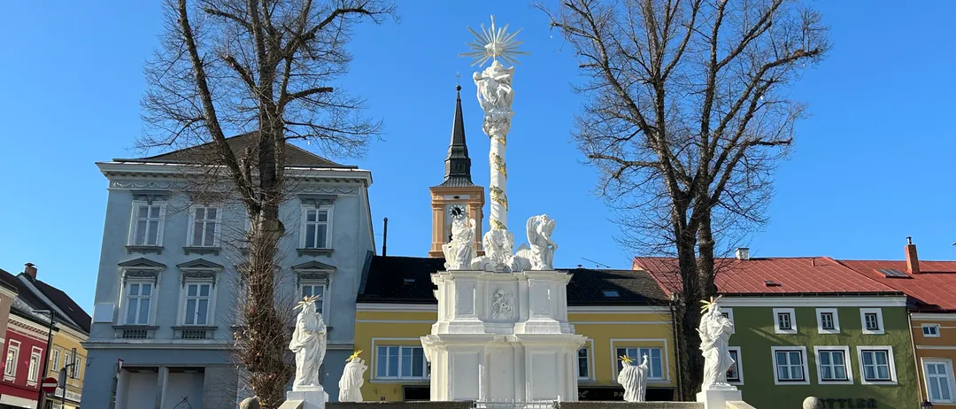 Eine weiße Statue mit einem Sonnenaufgang oben steht vor einem Gebäude mit einem Glockenturm, umgeben von Bäumen und Treppen.