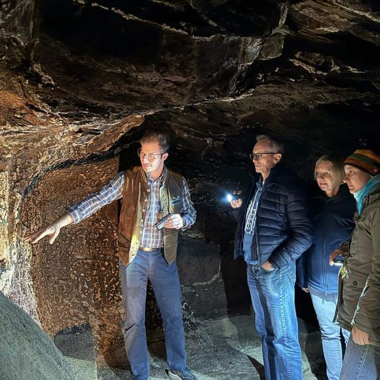 Ein Mann in einer braunen Weste gestikuliert und hält eine Taschenlampe in einer Höhle. Neben ihm stehen zwei Männer und eine Frau, ebenfalls mit Jacken bekleidet, alle schauen auf die Wand.