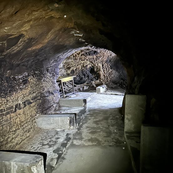 Innenansicht einer Höhle mit einem schmalen Betonweg. Die Höhlenwände bestehen aus Stein, und es gibt einen Holztisch mit einigen Trümmern. Die Höhlendecke hat ein kleines Loch.