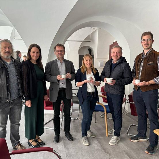 Sechs Personen stehen in einem Raum, lächeln und halten Tassen. Drei Männer und drei Frauen. Der Raum hat weiße Wände und eine Decke mit einem Bogendesign.